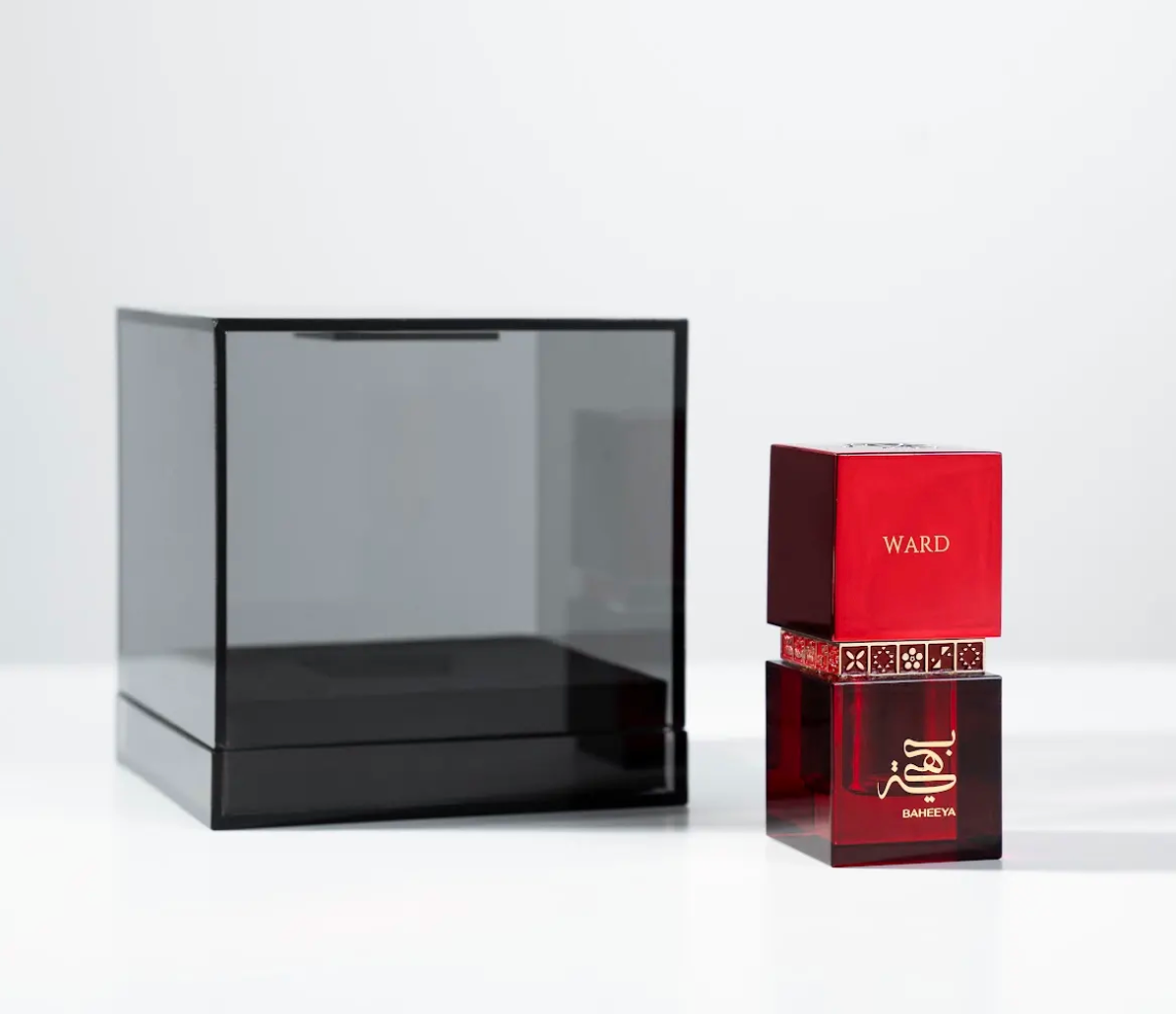 A dark translucent display case stands beside a small perfume bottle and its red box on a clean white studio background. The bottle and packaging show the brand name BAHEEYA and the fragrance name WARD.