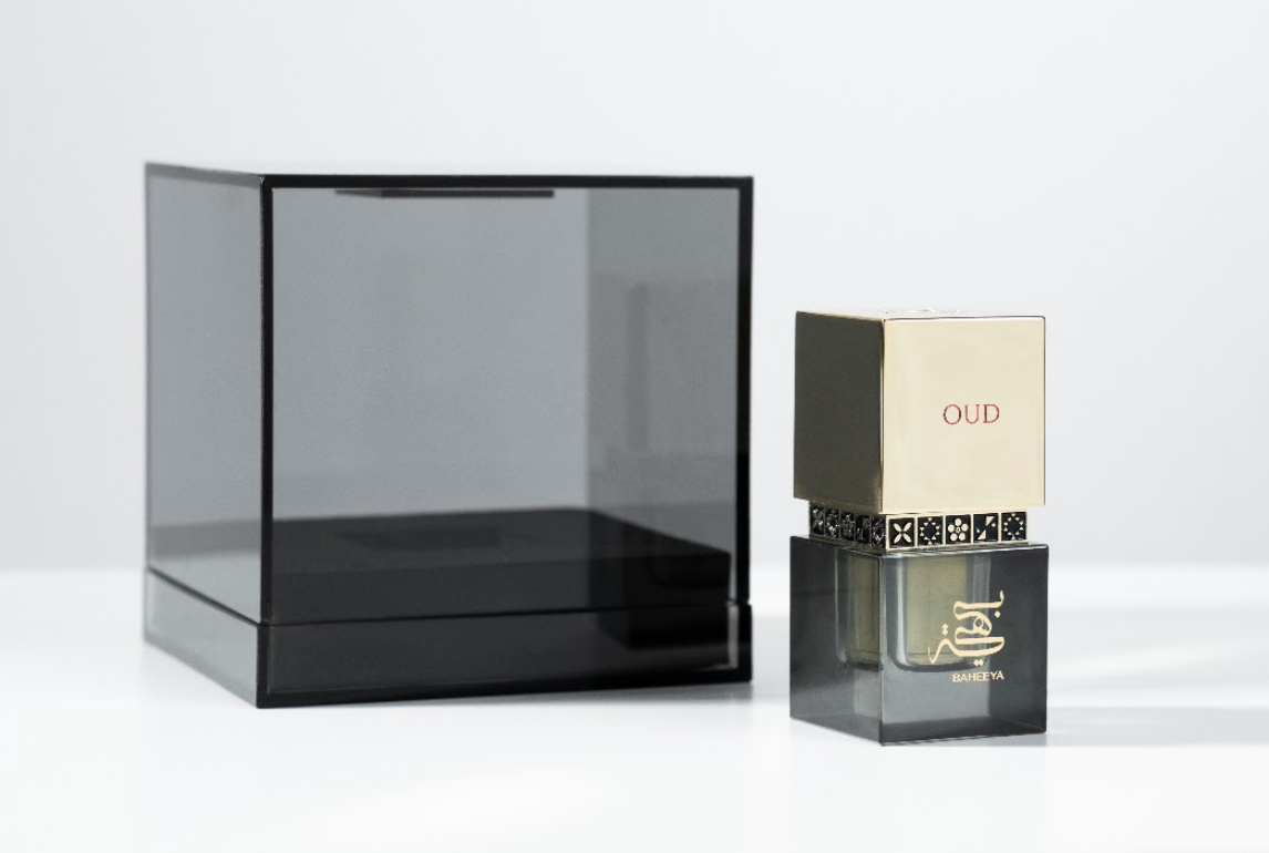 A perfume bottle with a gold cap and black patterned band next to a large transparent smoky black display box on a clean white background.