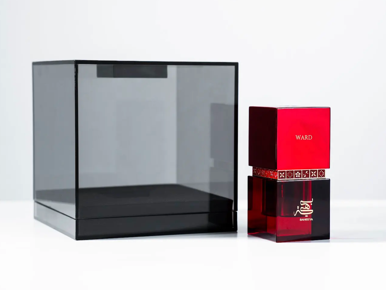 A red and black perfume bottle next to a large transparent black display box on a clean white background. The bottle features a red top section with the brand name 'WARD' and a patterned band, and a dark red translucent lower section with gold lettering.